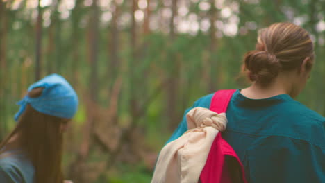 back view of young hikers hanging bags over their shoulders in a serene forest, woman in green shirt holds red bag with cloth wrap, while her companion, wearing a blue bandana, stands nearby