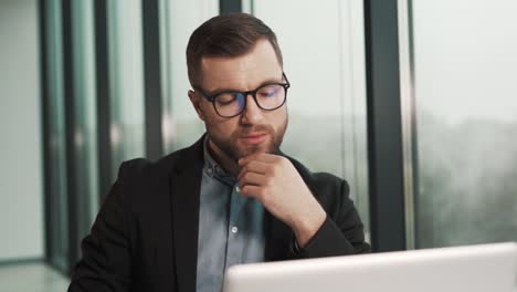 Concentrated-man-in-a-jacket-is-thinking-while-sitting-near-the-panoramic-window-at-the-laptop