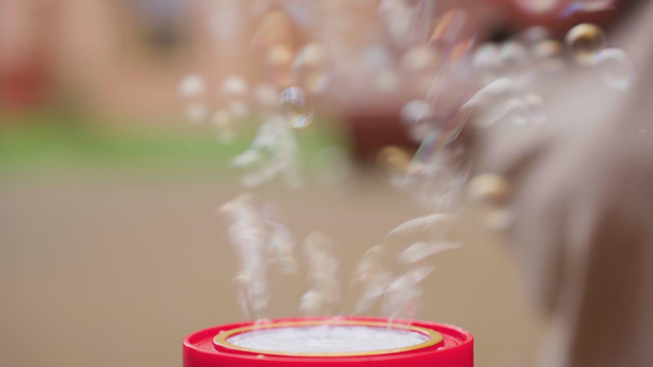 Close up of red bubble machine releasing clusters of delicate floating soap bubbles, blurred figure playing in background, soft focus captures movement