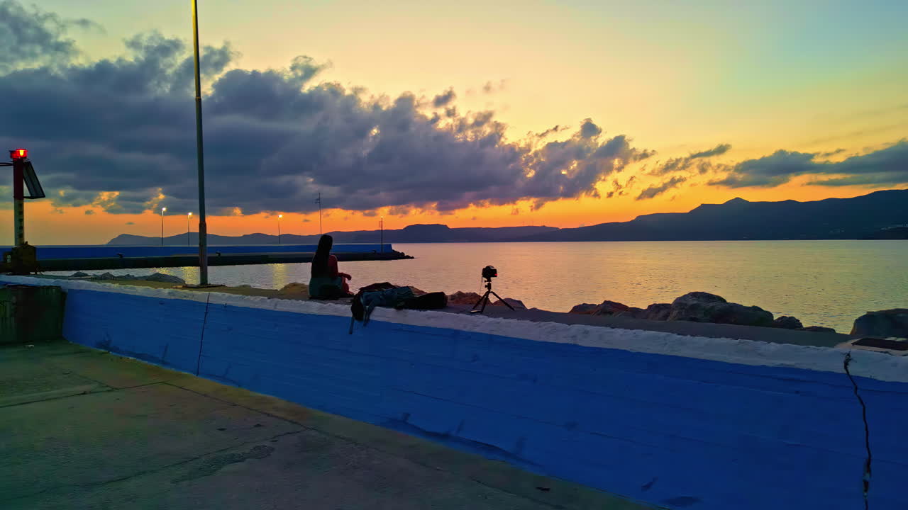 Drone fly sunset landscape, woman looking at the sky in Sitia Town in Crete, Greece, calm beach, water reflected, orange glow sky