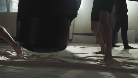 fighters exercise kicks on punching bags in gym closeup. barefoot kick-boxers do intense workout with heavy equipment in fighting club slow motion