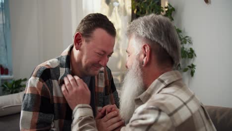 A-happy-brunette-man-in-a-plaid-shirt-holds-hands-with-his-elderly-boyfriend-with-gray-hair-and-a-lush-gray-beard.-Two-men-lean-their-foreheads-against-each-other-while-sitting-on-a-sofa-in-a-modern-apartment