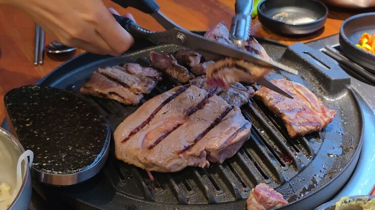 Potongan daging sapi BBQ Korea yang berair dipanggang di atas piring panggangan panas, dibalik dengan penjepit, dikelilingi oleh lauk pauk