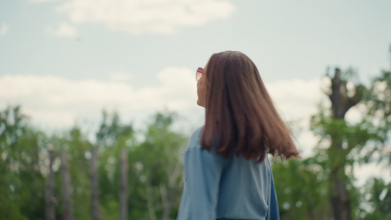 Mujer feliz al aire libre, mujer sonriente con el pelo ondeando al viento en un parque, alegre mujer en camisa vaquera disfrutando del aire fresco, mujer radiante con gafas de sol y el pelo alborotado por el viento en un animado evento en el parque