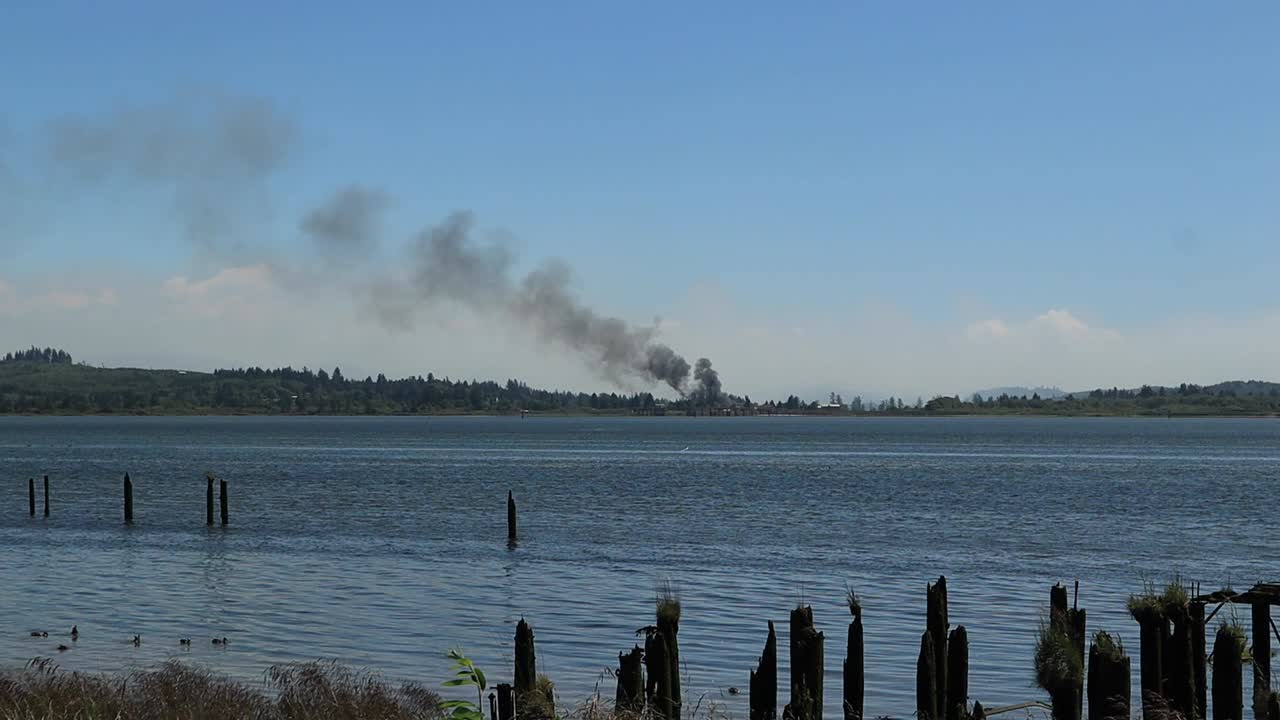утки плавают в воде, когда дым от пожара в лагере рилеа на берегу залива янгс в штате орегон, глядя на пилоны со старого причала, когда дым поднимается вверх и влево.