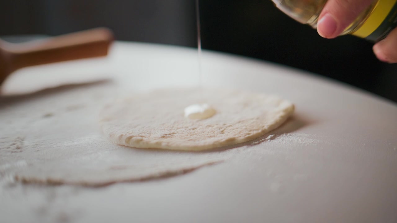 Hand sprinkles oil onto a flat dough circle. Dough rests on a floured white surface. Rolling pin lies nearby, unused. Scene shows calm, focused cooking preparation. Ideal for food blogs, recipe videos