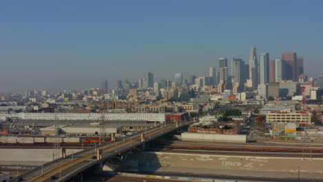 AERIAL:-LA-River,-Train-Tracks-with-Downtown-Los-Angeles-in-Background,-Sunlight