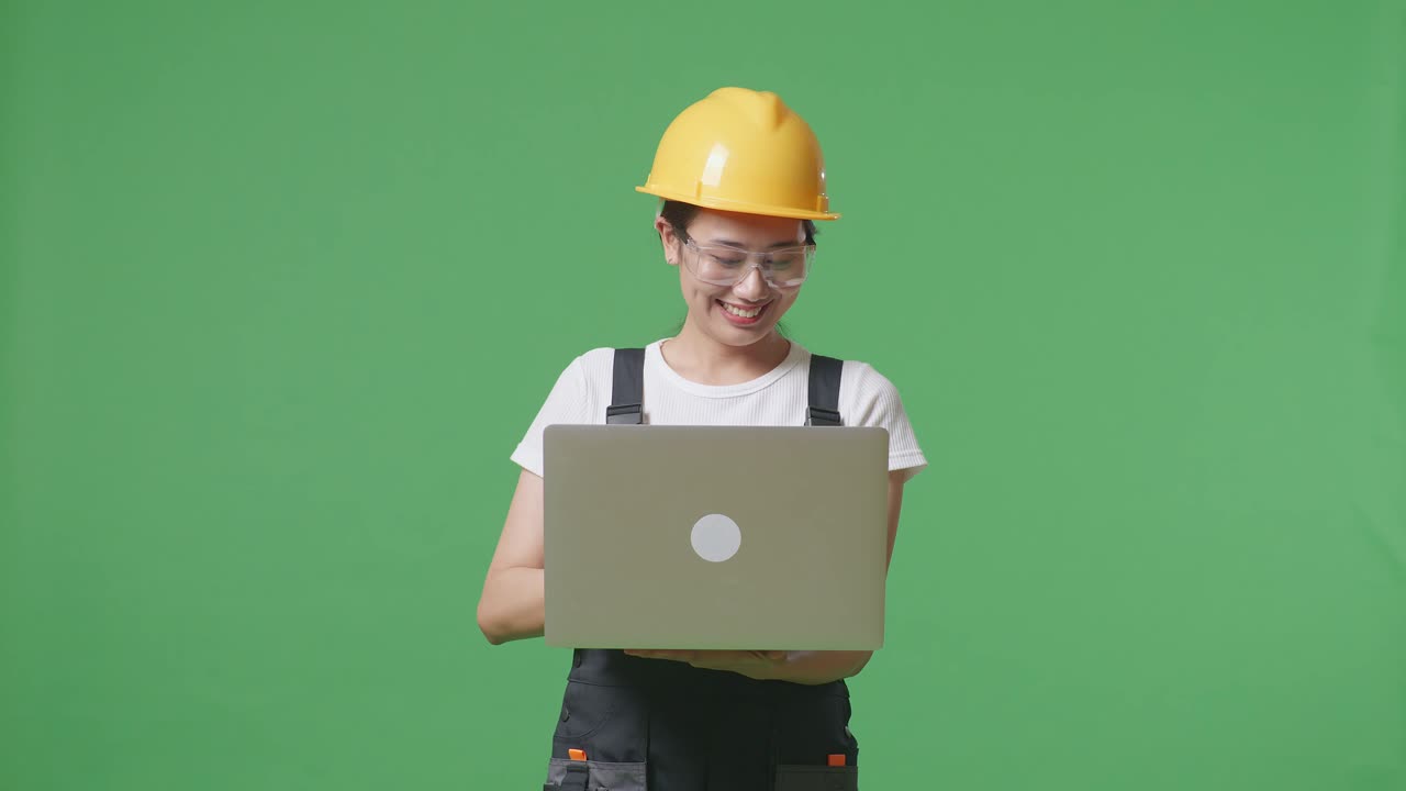 녹색 스크린 배경 스튜디오에 서있는 동안 노트북을 사용하여 보호 안경과 안전 헬을 착용 한 아시아 여성 노동자