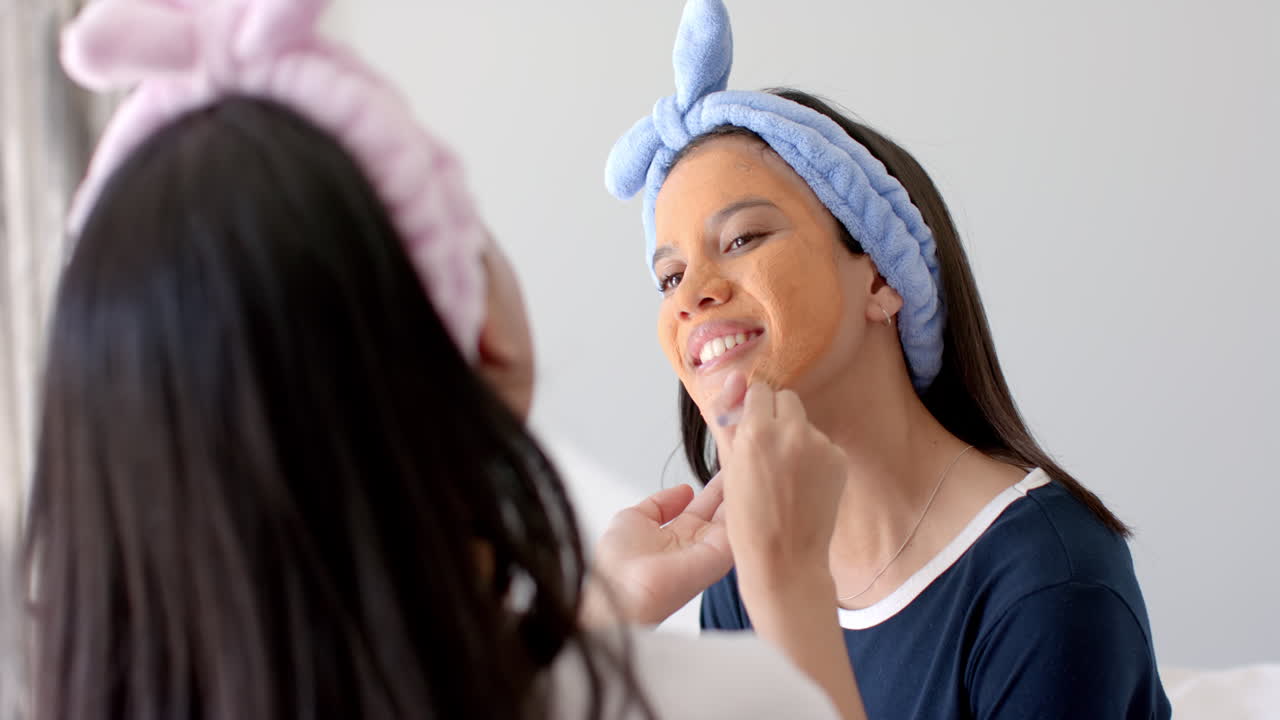 Two biracial teenage girl friends applying face mask at home, one with a blue headband