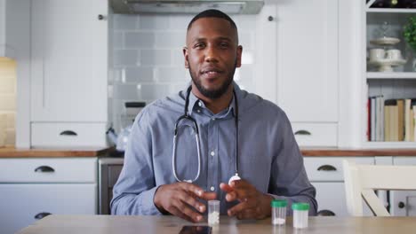 Portrait-of-african-american-doctor-holding-medication-container-talking-while-having-a-video-call-a