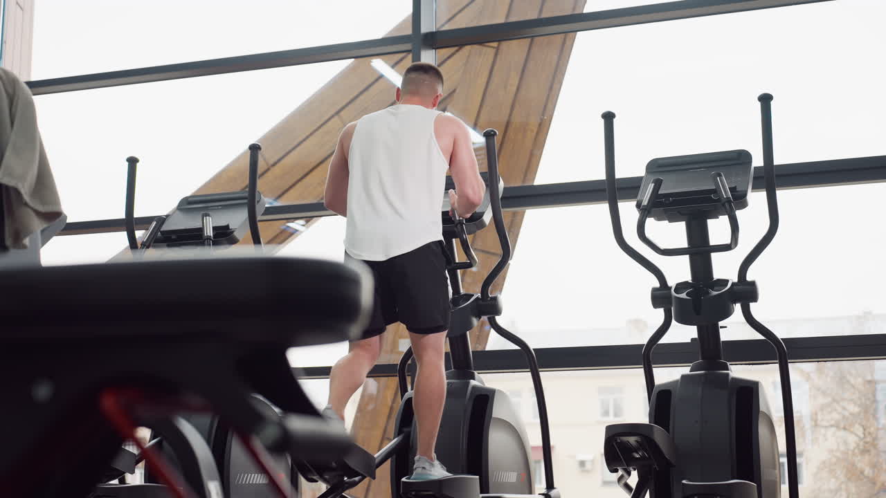 Fitness enthusiast concentrating on elliptical machine workout by angled windows in modern gym, stepping on pedals and adjusting console while glancing around, towel hanging on chair