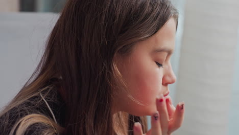 Side profile of young girl with long brown hair eating chips indoors, background includes soft furnishings and daylight from nearby window