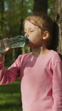 tired little girl drinks water leaning on old tree trunk in green park closeup. stay hydrated to avoid heat stroke. refreshing drink in hot weather