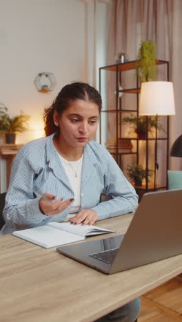 young woman writing down notes while attending online office meeting on laptop at home office desk
