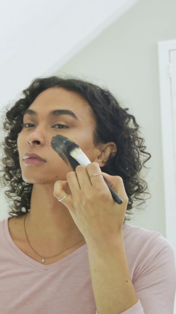 Vertical video: Seeing mirror nonbinary adult applying dark mask with brush at vanity for skincare