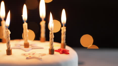 close up of lit candles on a white, decorated birthday cake, bokeh lights in the background