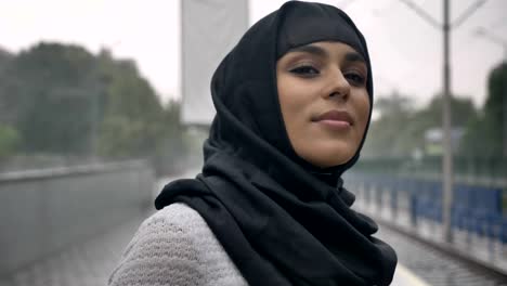 young muslim woman in hijab is waiting for train on railway station, raining, religion concept, urban concept. weather concept