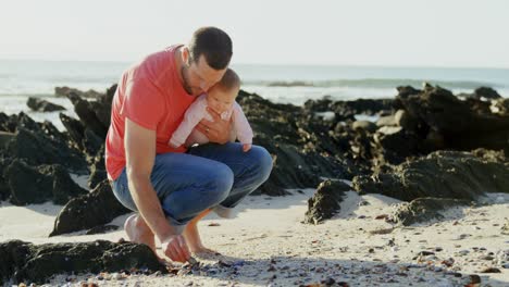 Vorderansicht-Eines-Kaukasischen-Vaters-Mittleren-Alters,-Der-Seinen-Sohn-Hält-Und-An-Einem-Sonnigen-Tag-Am-Strand-Kauert-4k