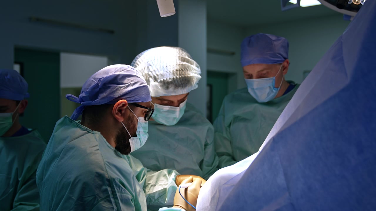 Tre chirurghi uomini circondano il tavolo operatorio in sala operatoria. Un medico barbuto usa uno strumento elettrico. Intervento chirurgico per la cura di un trauma.