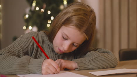 Close-Up-View-Of-A-Girl-In-Green-Sweater-Writing-A-Letter-And-Thinking-Of-Wishes-Sitting-At-A-Table-In-A-Room-Decorated-With-A-Christmas-Tree-1