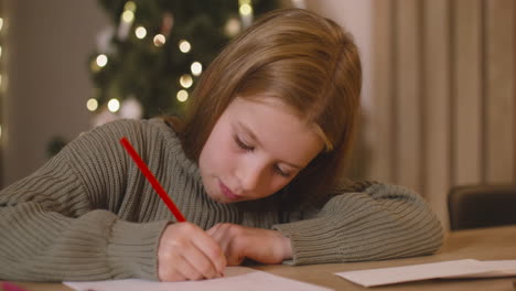 close up view of a girl in green sweater writing a letter and thinking of wishes sitting at a table in a room decorated with a christmas tree 1