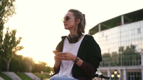 Low-angle-footage-of-a-pensive-girl-sitting-outdoors-on-the-street-or-park-with-her-smartphone-in-hands.-Profile-of-a-thoughtful