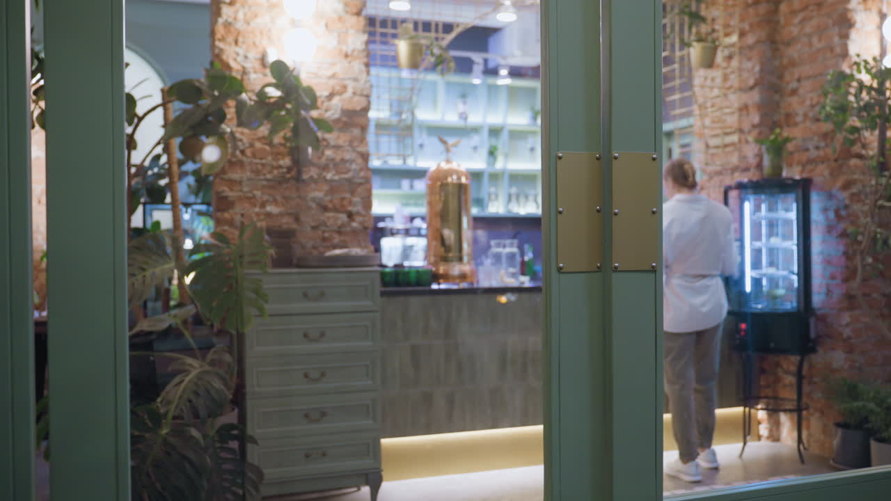 Back view of woman wearing white top, gray joggers, and sneakers as she retrieves item from fridge in cozy brick-walled cafeteria with indoor plants, wooden elements, and warm ambient lighting