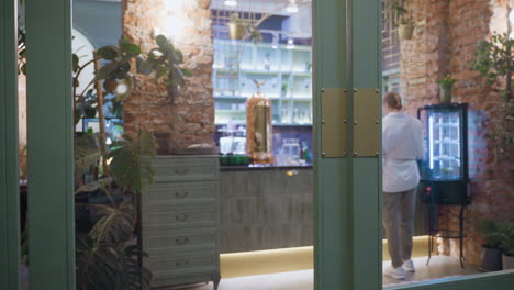 back view of woman wearing white top, gray joggers, and sneakers as she retrieves item from fridge in cozy brick-walled cafeteria with indoor plants, wooden elements, and warm ambient lighting