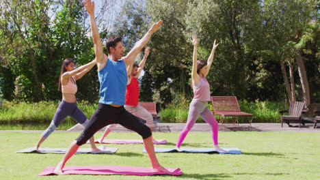 Diverse-group-practicing-yoga-with-male-instructor,-standing-on-mats-in-sunny-park