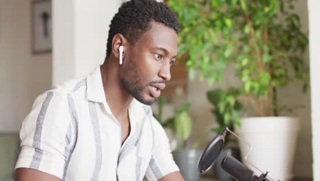 happy african american man sitting at table in kitchen, using tablet and making vlog