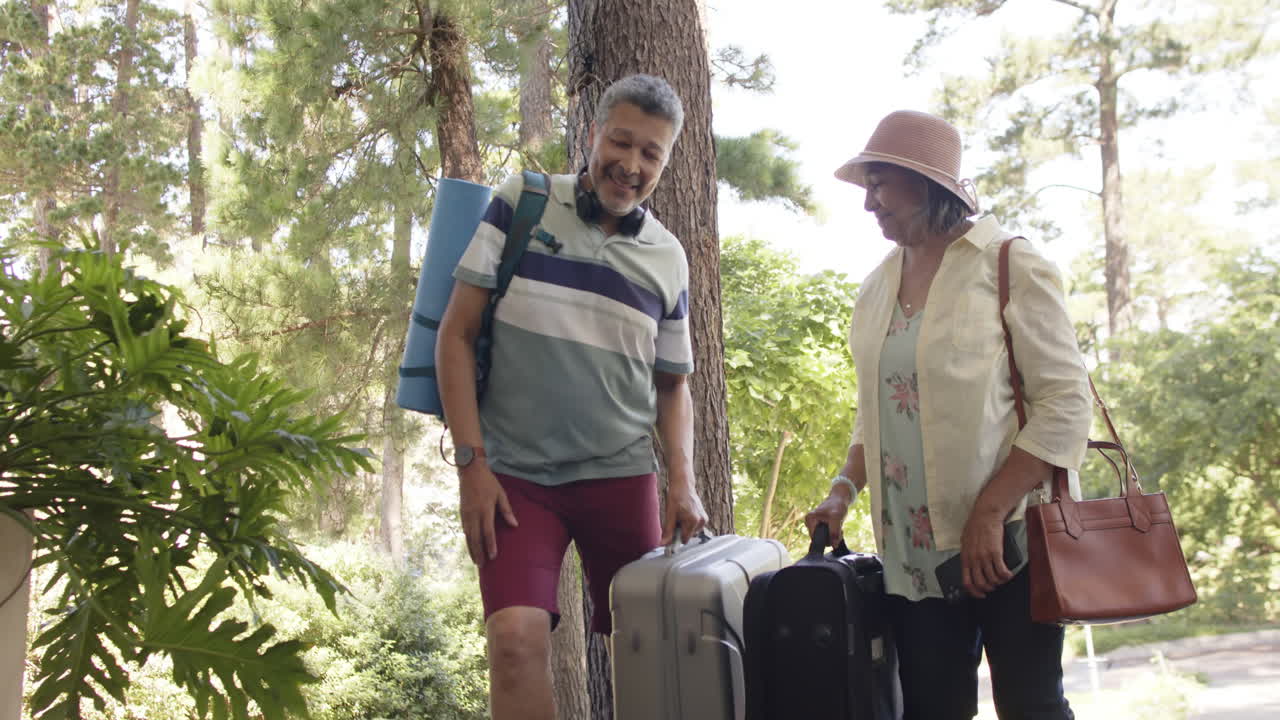 feliz pareja de ancianos diversos caminando con el equipaje a una casa al aire libre soleado