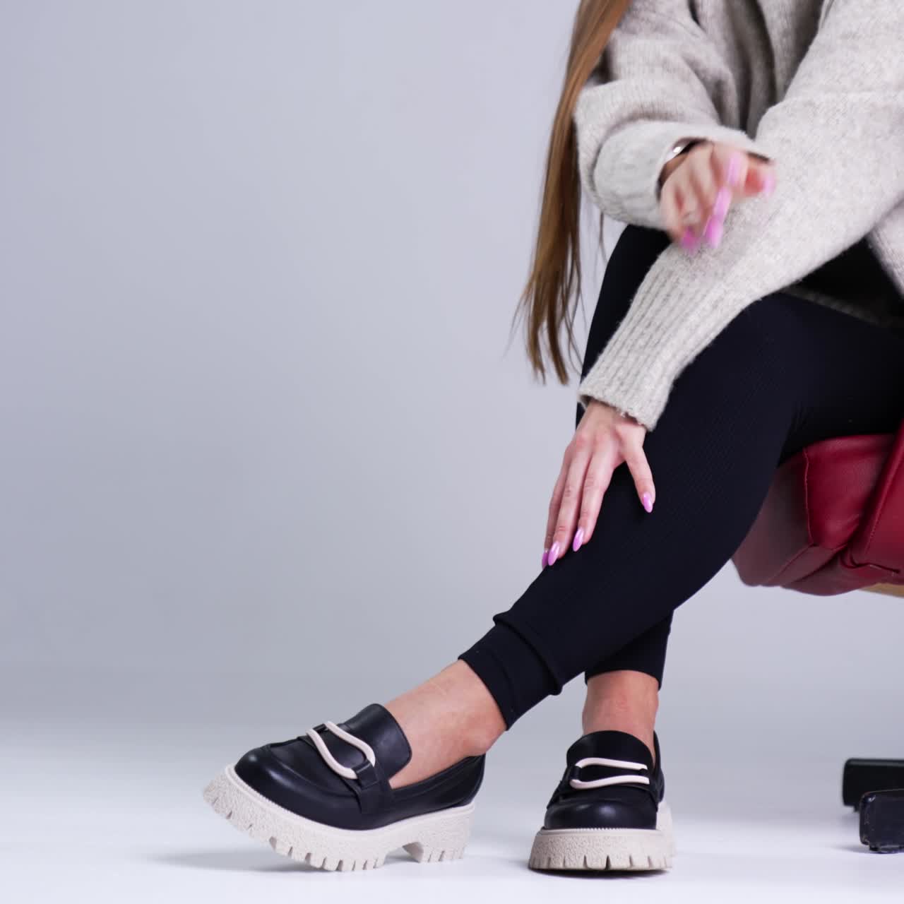 Female sitting in a red chair stretching hand to her shoes. Modern stylish footwear demonstration in the studio. White backdrop