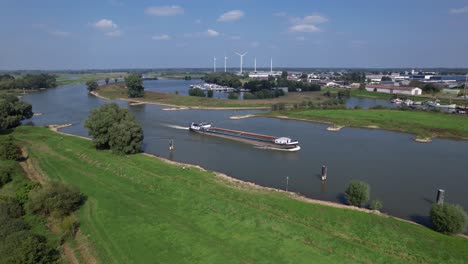 Rückwärtsbewegung-In-Der-Luft-Nach-Einem-Leeren-Frachtschiff,-Das-Auf-Der-Ijssel-Bei-Zutphen-Vorbeifährt,-Mit-Überschwemmungsgebieten-Und-Windkraftanlagen-Im-Hintergrund