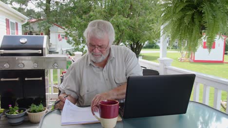 a senior man sits at an outdoor table, using a laptop while sipping coffee. the setting features a grill, plants, and a serene backyard, creating a perfect morning work atmosphere