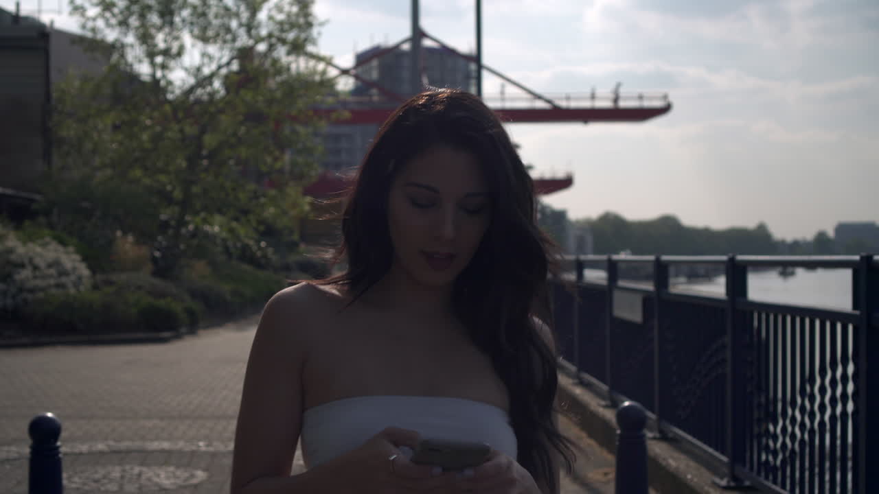 캐주얼 상의를 입은 자신감 있고 매력적인 젊은 브루네트 소녀는 휴대폰을 사용하고 문자 메시지에 행복하게 미소를 지으며 주위를 둘러봅니다.