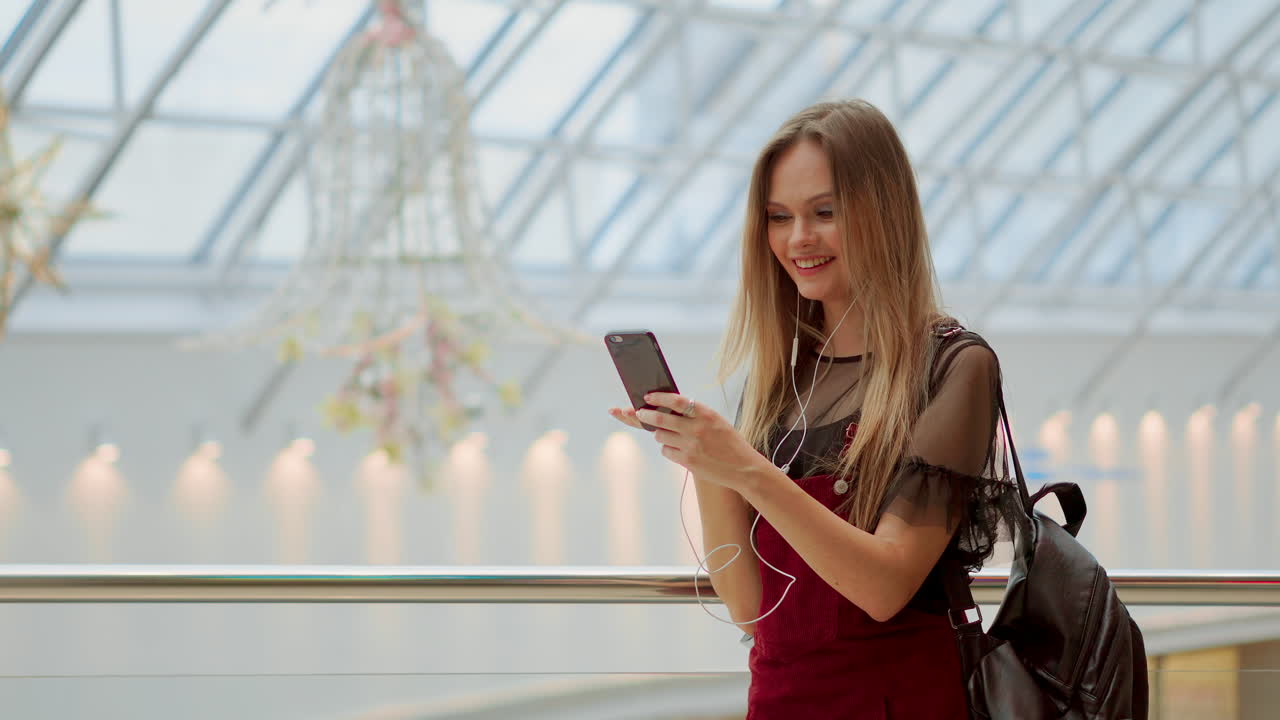 Girl with a backpack talking on video coll through headphones and a mobile phone at the airport. posing for selfie on smartphone camera, hipster girl enjoying radio broadcast making photo for picture in social networks on mobile.