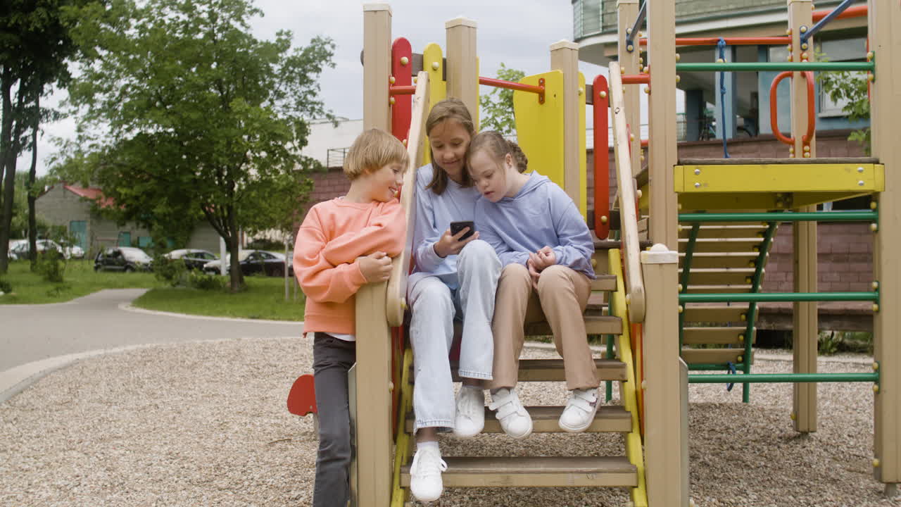 Little girl with down syndrome and her friends laughing and watching a video on smartphone in the park on a windy day