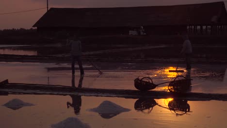 Workers-prepare-the-salt-fields-for-harvest,-woven-baskets-ready,-silhouetted-in-the-glistening-evening-sun