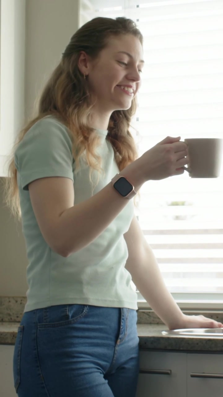 Video vertical: Mujer con camiseta verde reaccionando a las persianas, sosteniendo una taza y tocando un reloj inteligente en la cocina.