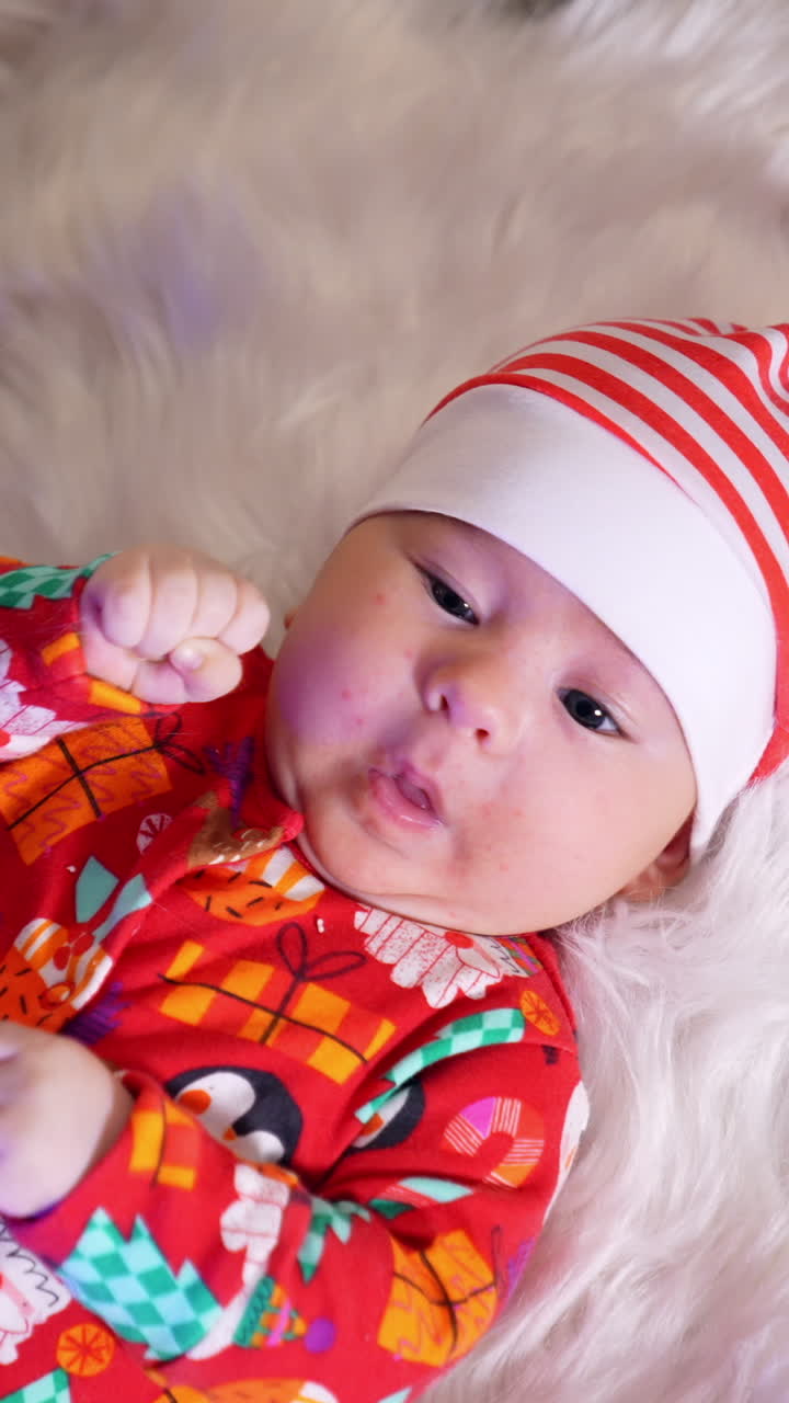 Serious little baby boy in red and white hat lies on white fluffy plaid. Funny kid near the decorated Christmas tree. View from above. Vertical video