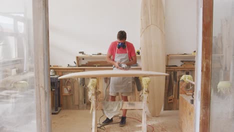 Caucasian-male-surfboard-maker-wearing-a-breathing-face-mask-and-shaping-a-wooden-surfboard-