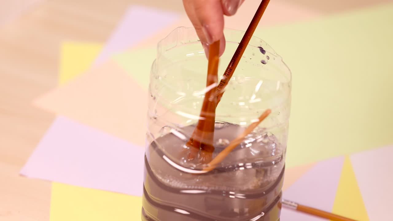 Hand cleans paintbrushes in water-filled plastic container, bright studio lighting, overhead camera angle