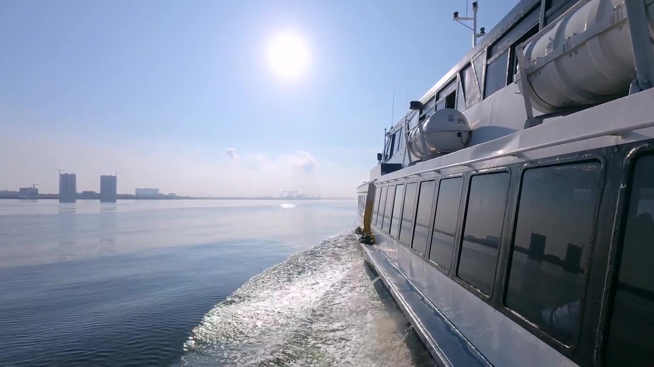 Eine Echtzeit-Seitenansicht einer Fähre mit 60 Bildern pro Sekunde, die die Heckwelle und Sonnenreflexion zeigt, mit einer entfernten Stadtsilhouette am Horizont.