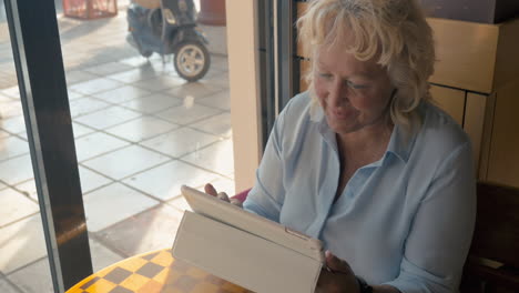 mujer madura sentada en un café y disfruta de su tableta fuera de la ventana el día soleado de verano