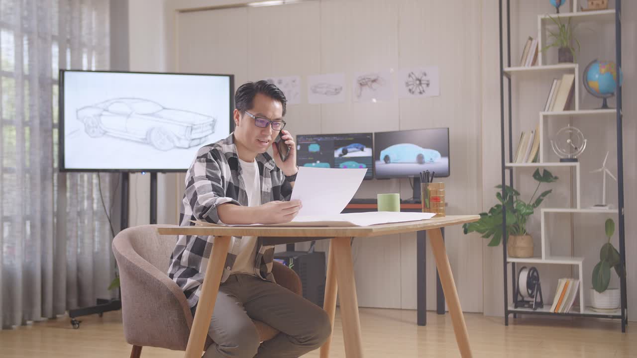 아시안 남성은 tv와 컴퓨터를 가진 스튜디오에서 테이블에서 작업하는 동안 스케치 및 스마트폰에서 이야기하는 자동차 디자인을보고 3d 전기 자동차 모델을 표시합니다.