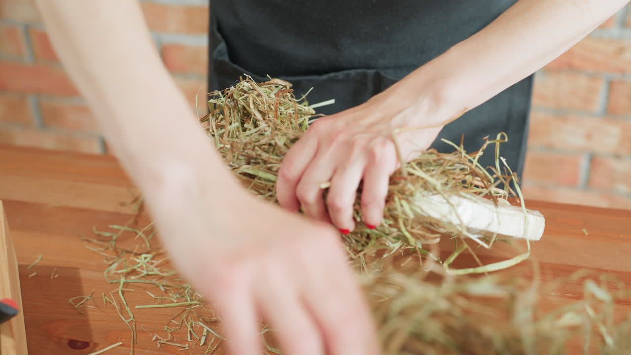 Florist arranges dried grass onto circular base using hands with red nails, preparing handmade wreath in creative workspace, natural materials and artistic floral craft process in rustic design project