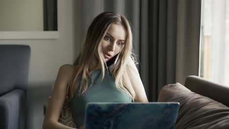 blonde woman sitting on sofa with laptop on knees and talking by smartphone