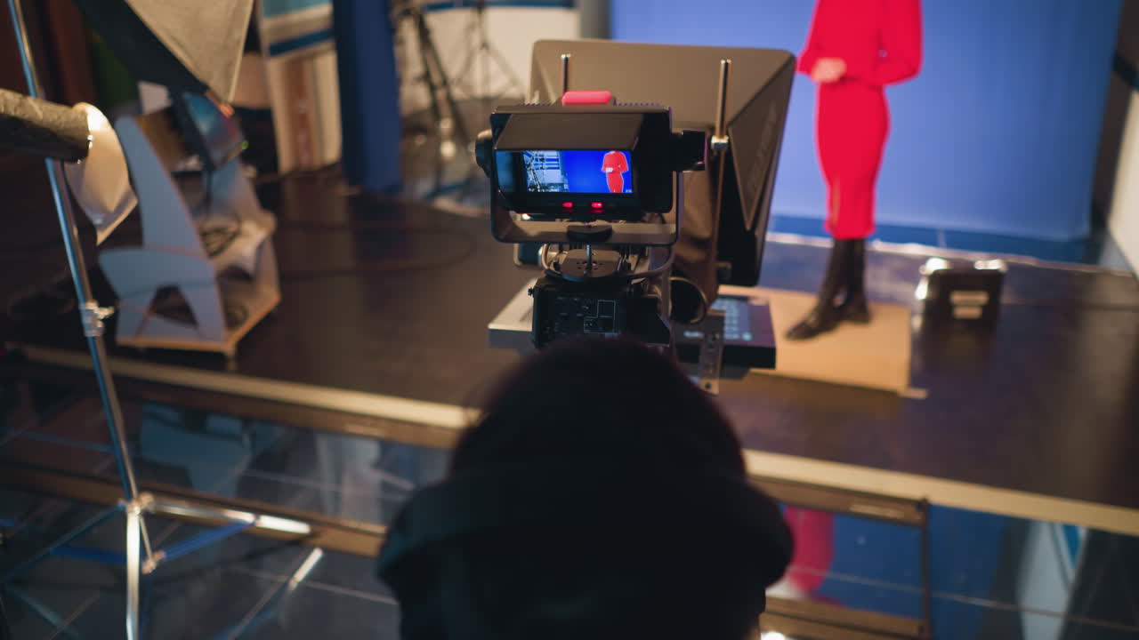 Cameraman operating studio camera with teleprompter and monitoring presenter in red dress on blue screen background during television production, weather forecast displayed on adjacent screen adds broadcasting context