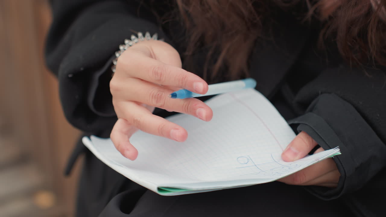 student filling notebook with blue pen during urban study session, closeup hands flipping pages, bracelet accent, brisk gestures for planning and scheduling, concentrated energy, breeze and campus