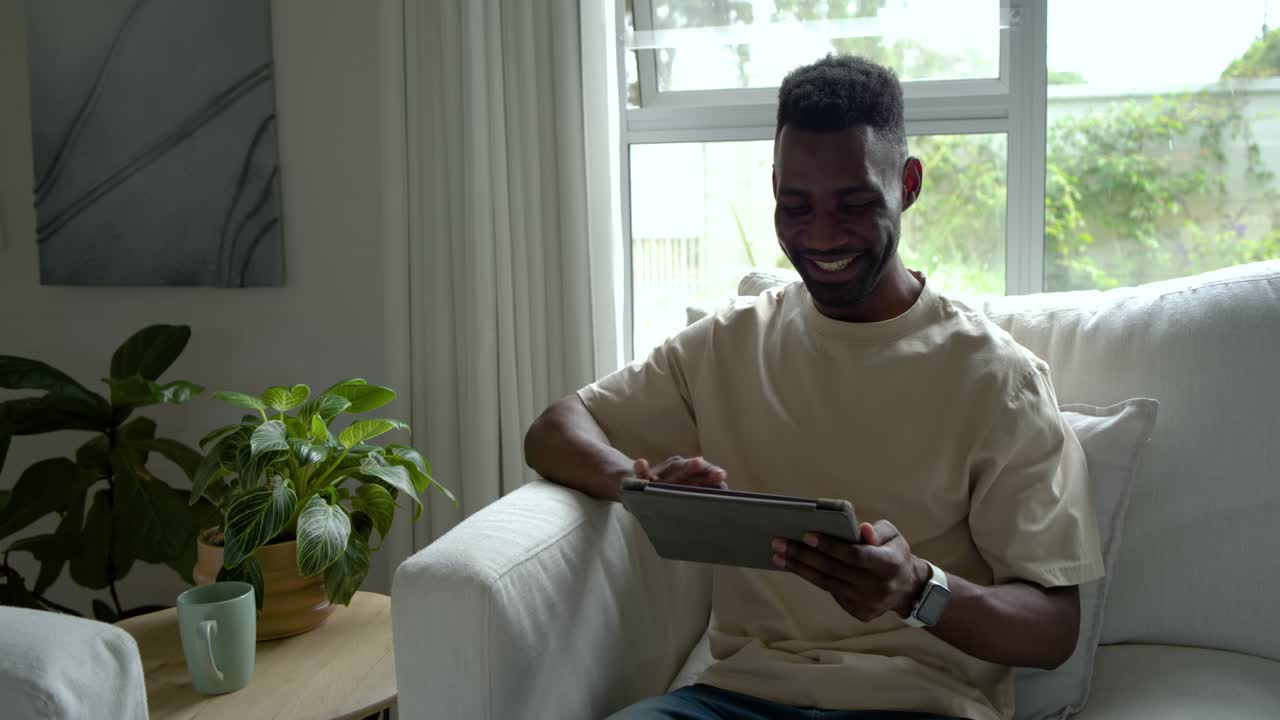 Adult African man smiling and tapping tablet, reacting to entertaining content in armchair at home